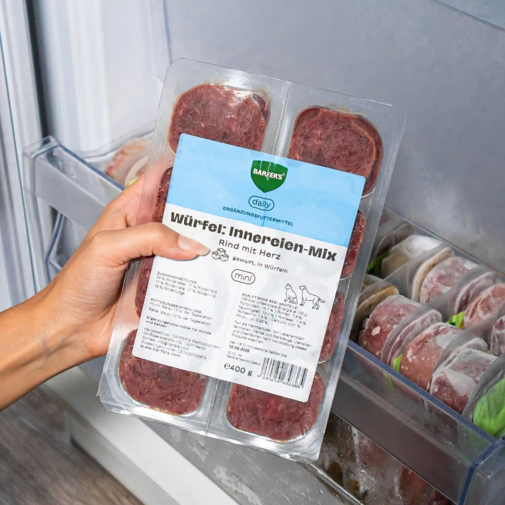 Hand hält eine 400g Verpackung BARFER’S Würfel Innereien-Mix Rind mit Herz für Hunde und Katzen vor ein geöffnetes Gefrierfach – sichtbar sind mehrere gefrorene Fleischwürfel