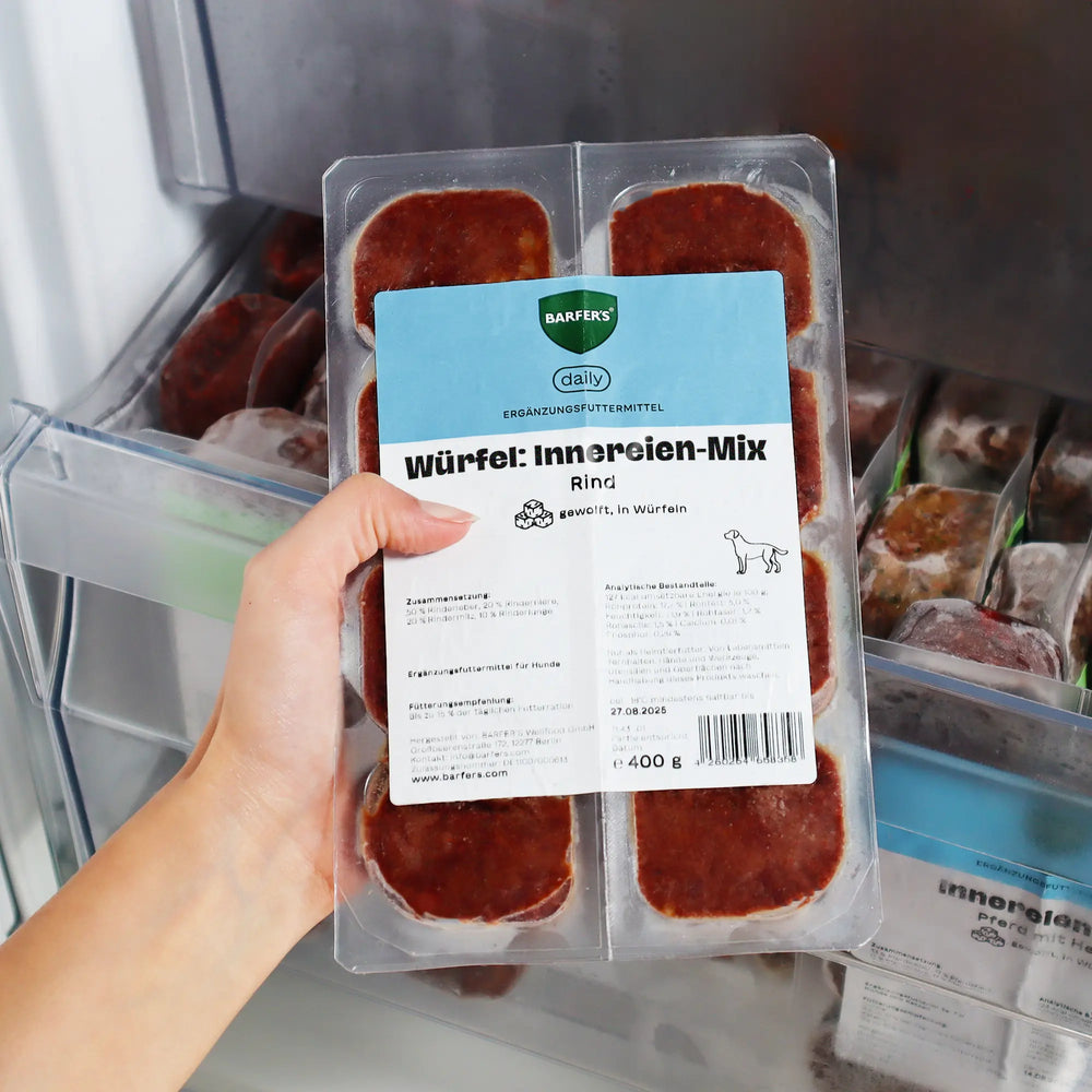 Hand hält 400g Verpackung vom BARFER’S Würfel Innereien-Mix Rind für Hunde mit 8 gefrorenen 50 g-Würfeln über ein geöffnetes Gefrierfach – Ergänzungsfuttermittel aus Rinderleber, Rinderniere, Rindermilz und Rinderlunge