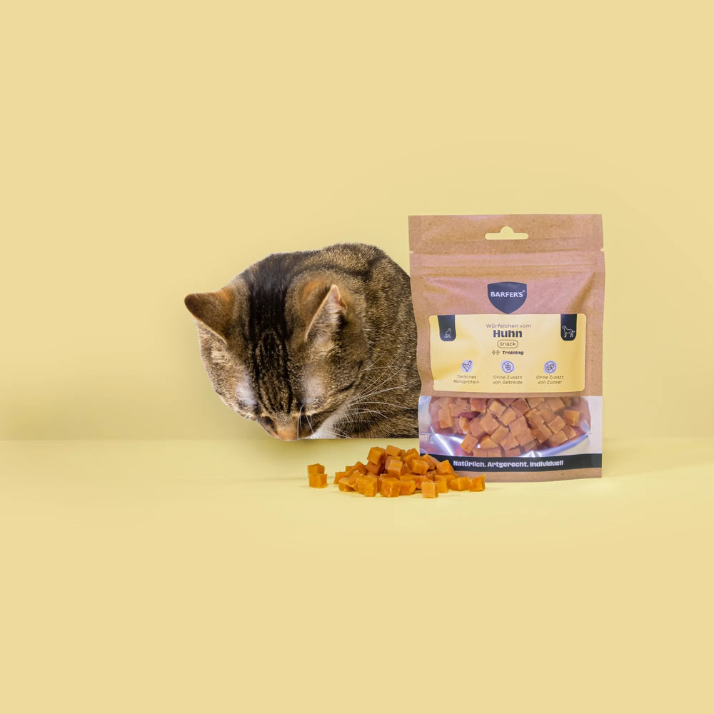 Eine schwarz-braun gestreifte Katze schaut Leckerli vom Huhn der Marke BARFER'S an, die neben einer vollen Verpackung "Würfelchen vom Huhn" liegen