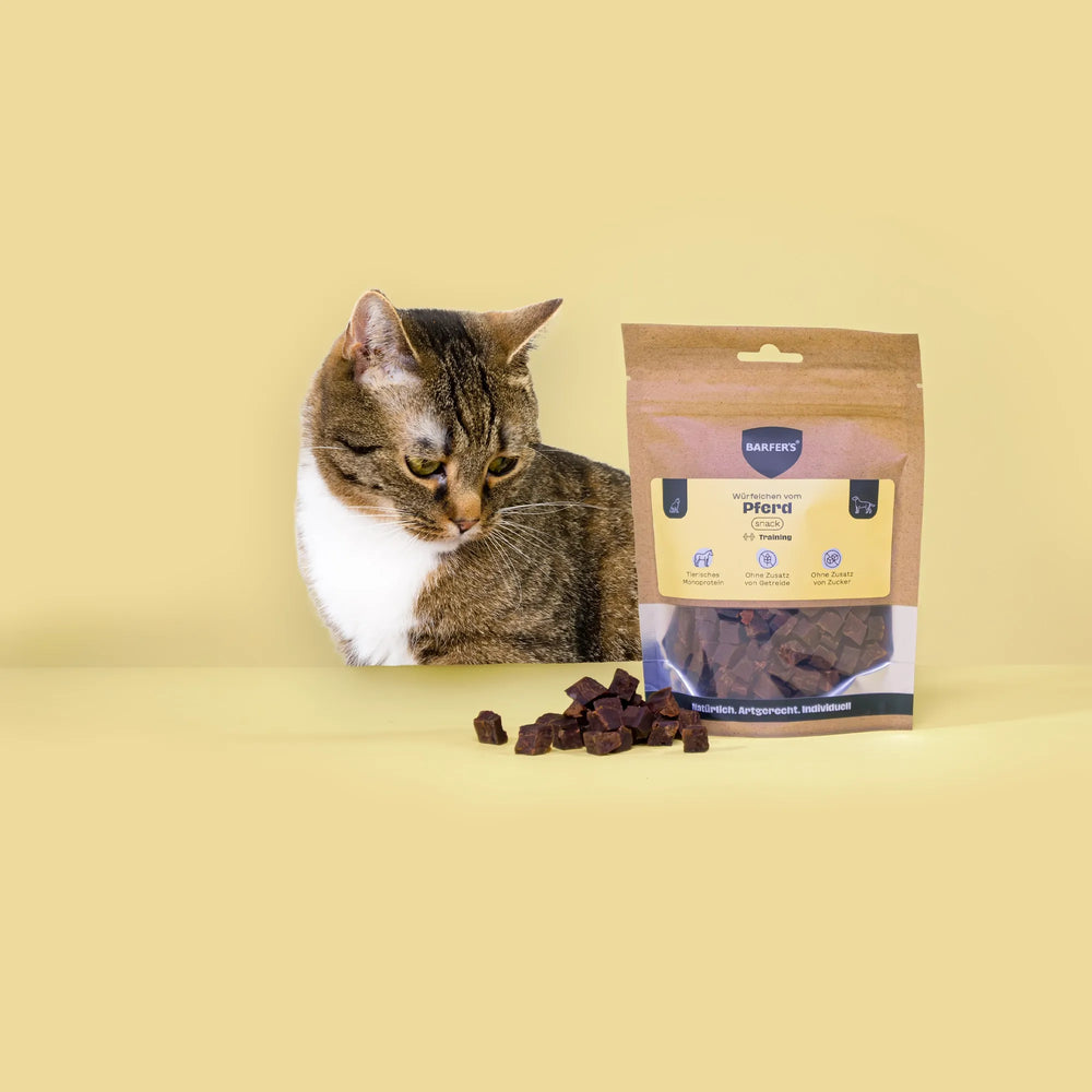Eine schwarz-grau gestreifte Katze schaut Leckerli vom Pferd der Marke BARFER'S an, die neben einer vollen Verpackung "Würfelchen vom Pferd" liegen