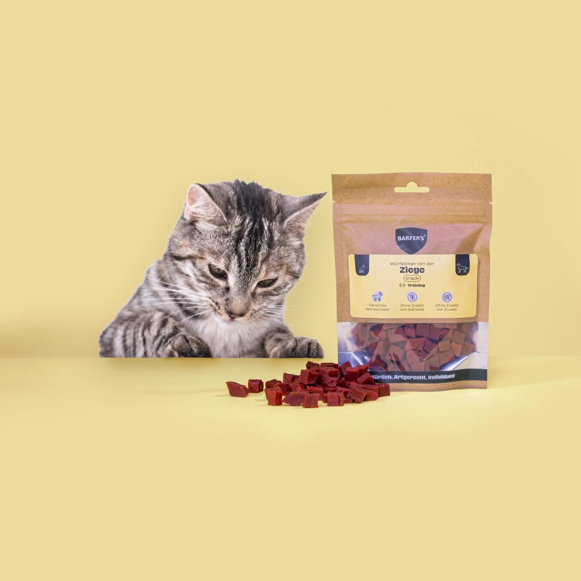 Ein schwarz-weiß getigerte Katze schaut neugierig auf Leckerli von der Ziege der Marke BARFER'S, die neben einer vollen Verpackung "Würfelchen von der Ziege" liegen