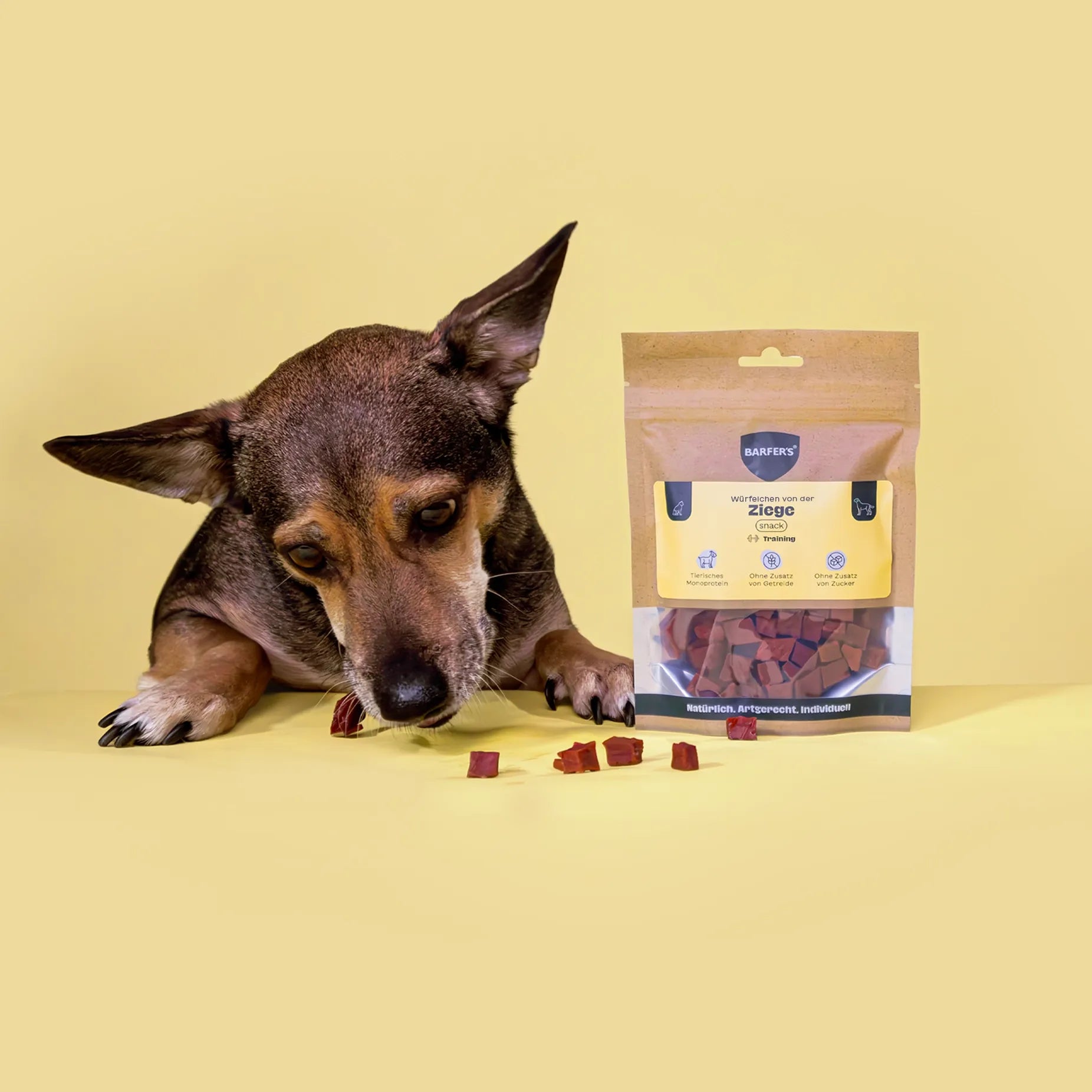 Ein schwarz-brauner Hund frisst gierig Leckerli von der Ziege der Marke BARFER'S, die neben einer vollen Verpackung "Würfelchen von der Ziege" liegen