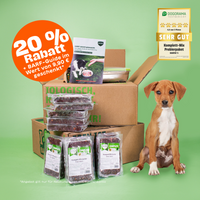 Junghund sitzt vor einem geöffneten Paket mit BARFER’S Komplett-Mix Junior Rind & Huhn, daneben BARF-Guide, Frischhaltedose, Rabatt-Badge mit 20 % und Hinweis auf Dogorama-Testurteil Sehr Gut (4,8 von 5 Pfoten)