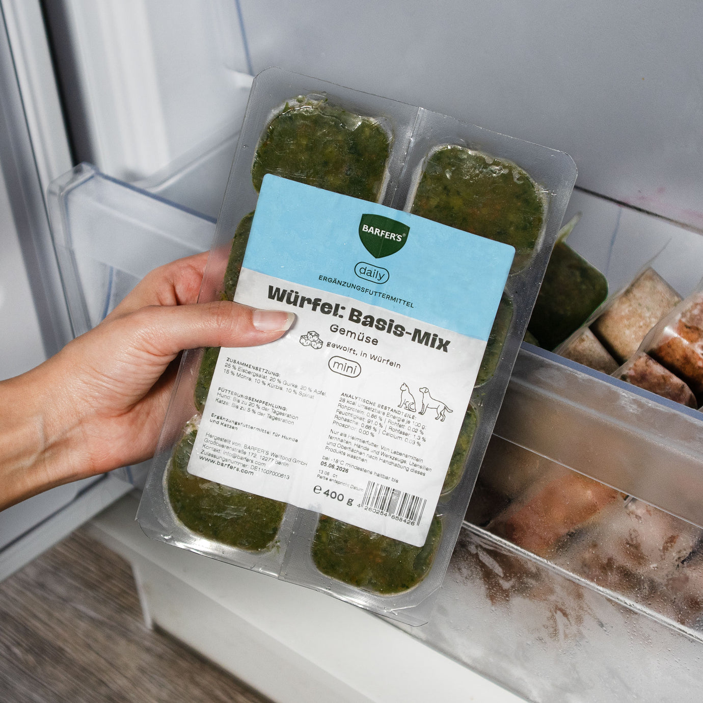 Hand hält 400g Verpackung vom BARFER’S Würfel Basis-Mix Gemüse mit 8 gefrorenen 50g-Würfeln über ein geöffnetes Gefrierfach – Ergänzungsfuttermittel aus Eisbergsalat, Gurke, Apfel, Möhre, Kürbis und Spinat