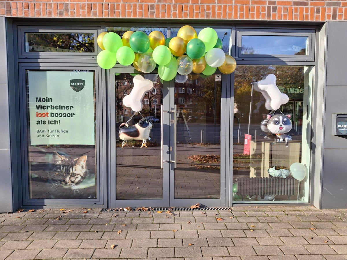 Außenansicht vom BARFER'S Store in Hamburg-Barmbek. Die 4 Glastüren sind zur Neueröffnung mit Luftballons geschmückt. Links außen hängt ein Plakat mit der Aufschrift "Mein Vierbeiner isst besser als ich! BARF für Hunde und Katzen."